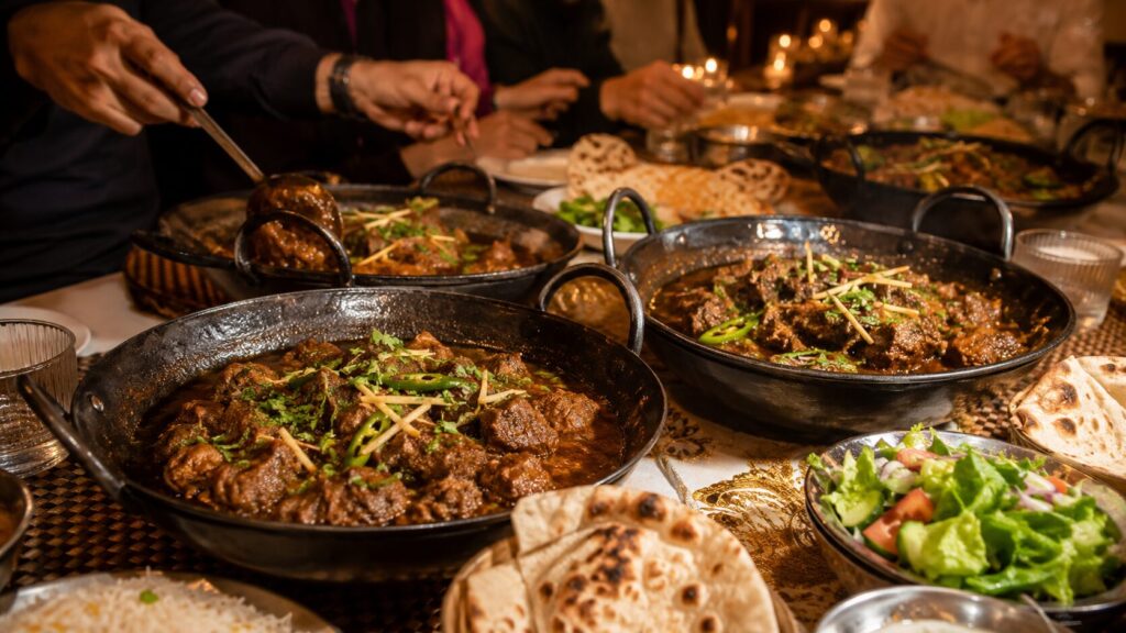 A festive dinner table set for an evening celebration, featuring several black iron karahi pans filled with steaming dumba karahi, garnished with fresh coriander and sliced green chilies. Warm naan, fluffy rice, and fresh salads surround the dishes, while guests' hands serve food. Soft string lights in the background and a shallow depth of field create a warm, inviting atmosphere.